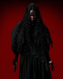 A haunting studio portrait of a woman in a black dress with red eyes against a red background.